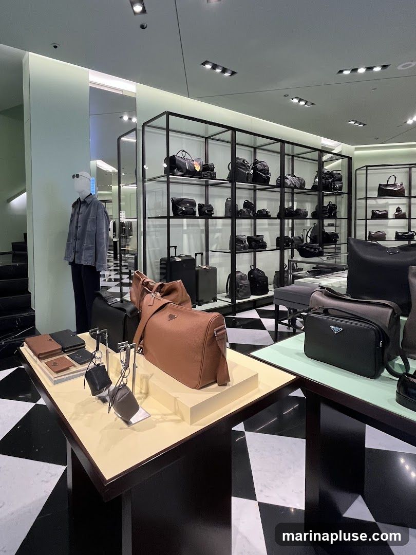 A well-lit interior of a luxury store showcasing a variety of handbags, luggage, and small leather goods. A mannequin dressed in a grey jacket stands to the left. The display features shelves filled with black bags and suitcases, while prominent tables in the foreground exhibit brown and black handbags, wallets, and keychains. The floor has a distinctive black and white checkered pattern.