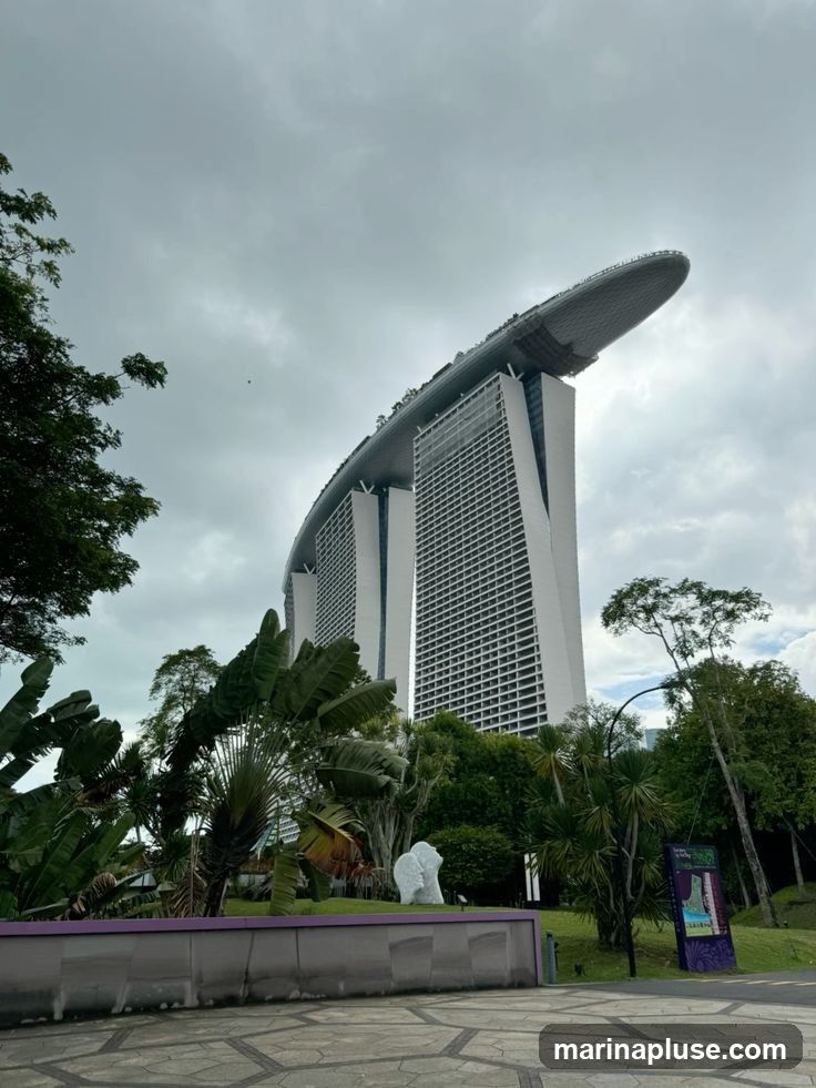 A low-angle shot of the Marina Bay Sands hotel in Singapore under a cloudy sky. Lush tropical foliage, including large palm leaves, is in the foreground. A stone wall with a purple trim runs along the bottom of the image. To the right, a colorful sign with illustrations and text is visible, likely an informational display or advertisement for the Gardens by the Bay.