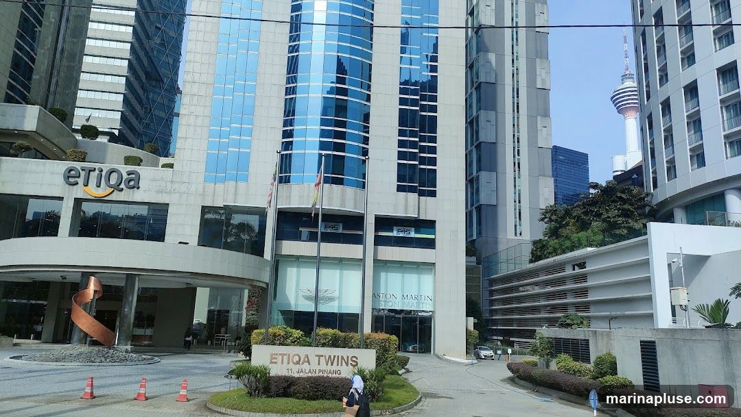A view of the Etiqa Twins building, a modern office complex with glass facades. The Etiqa logo is prominently displayed on the upper left side of the building. Below, the Aston Martin logo is visible on the ground floor. A sign at the entrance reads "ETIQA TWINS 11, JALAN PINANG". The Kuala Lumpur Tower can be seen in the distance.