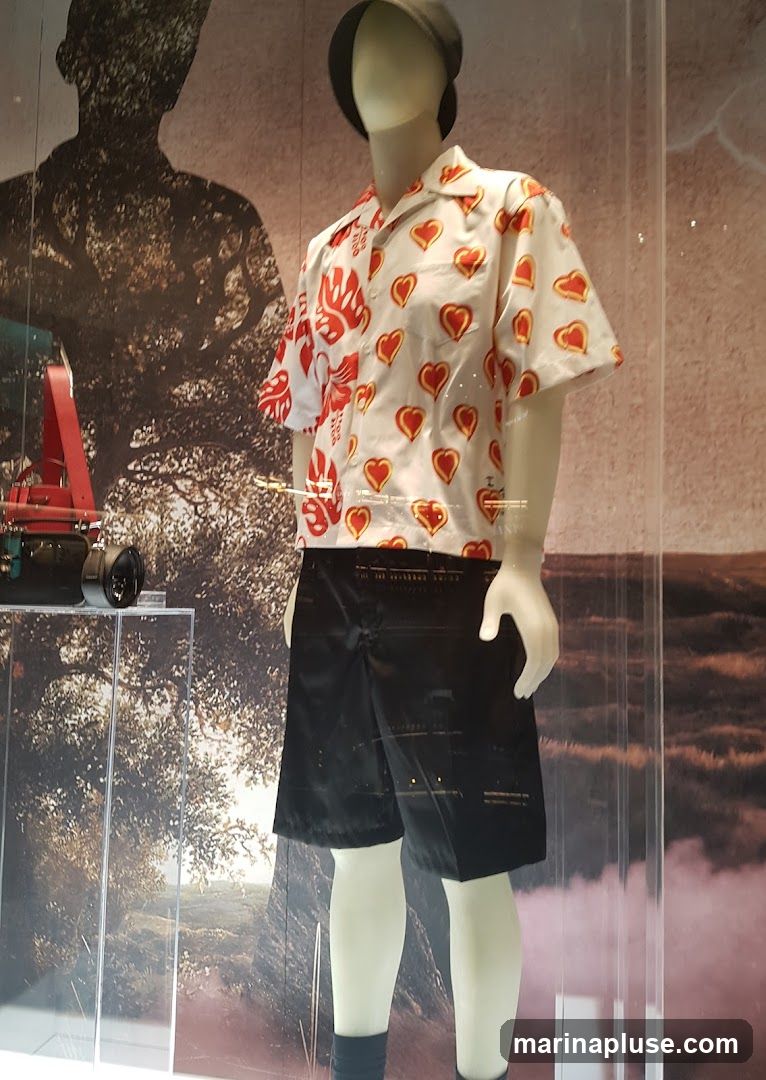 A mannequin is dressed in a white short-sleeved shirt with a red and yellow heart pattern and black shorts. The mannequin is wearing a dark cap and dark socks. The background of the display features a silhouette of a person against a tree and a landscape scene. To the left, a red strap and a black cylindrical object are visible on a clear shelf.