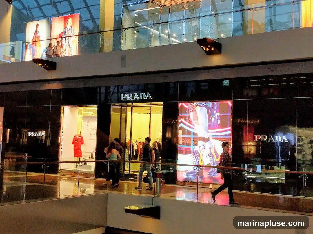 The exterior of a Prada store in a modern shopping mall. The facade is black with large glass windows and doors. The Prada logo is prominently displayed in white lettering above the entrance and on the glass. Inside, mannequins display clothing, and racks of garments are visible. Digital screens on the facade show fashion imagery. People are walking past the store.