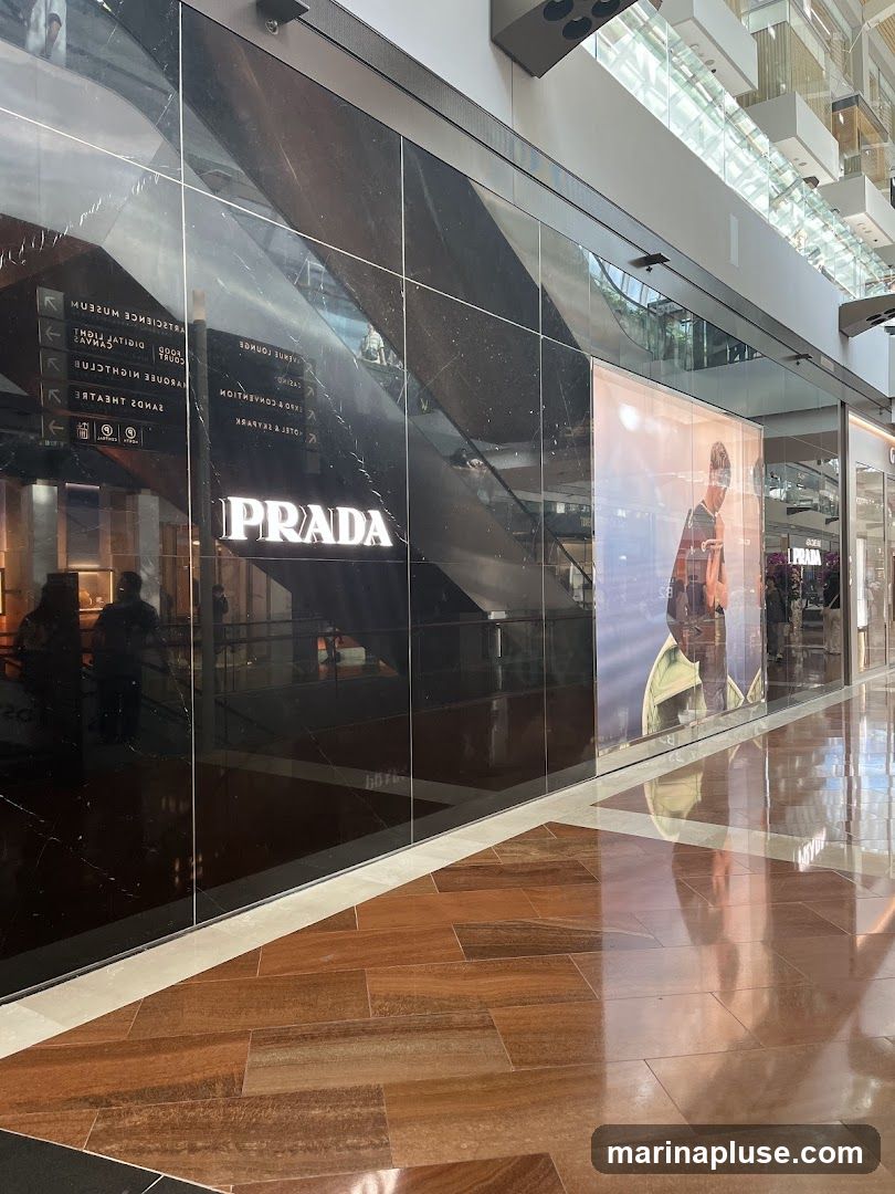 A sleek, modern storefront for Prada, featuring a black marble facade with the illuminated Prada logo. To the right of the logo, a large advertisement displays a fashion model. The floor is made of polished wood-like tiles, reflecting the interior lights and the store's facade. Signs indicating directions to various attractions like the ArtScience Museum and Sands Theatre are visible on the left side of the facade, reflected in the glass.