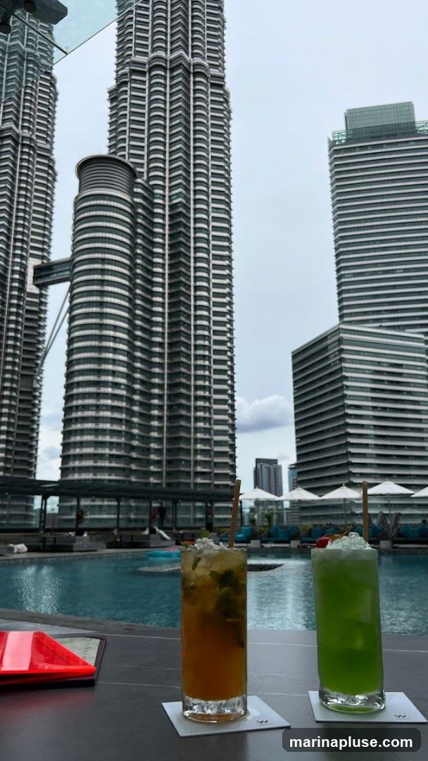 Two tall cocktails, one amber and one green, sit on a bar counter in front of a swimming pool. In the background, the iconic Petronas Twin Towers and other skyscrapers dominate the skyline under an overcast sky. Lounge chairs and umbrellas are visible around the pool area.