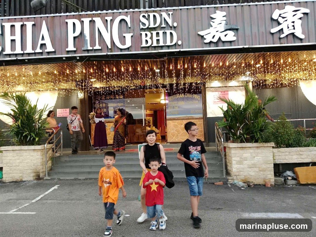 The exterior of a restaurant with a large sign that reads "CHIA PING SDN. BHD." and Chinese characters. The entrance is decorated with numerous strings of small lights. Several people are visible on the steps leading into the restaurant, and a family with children is walking in the foreground.