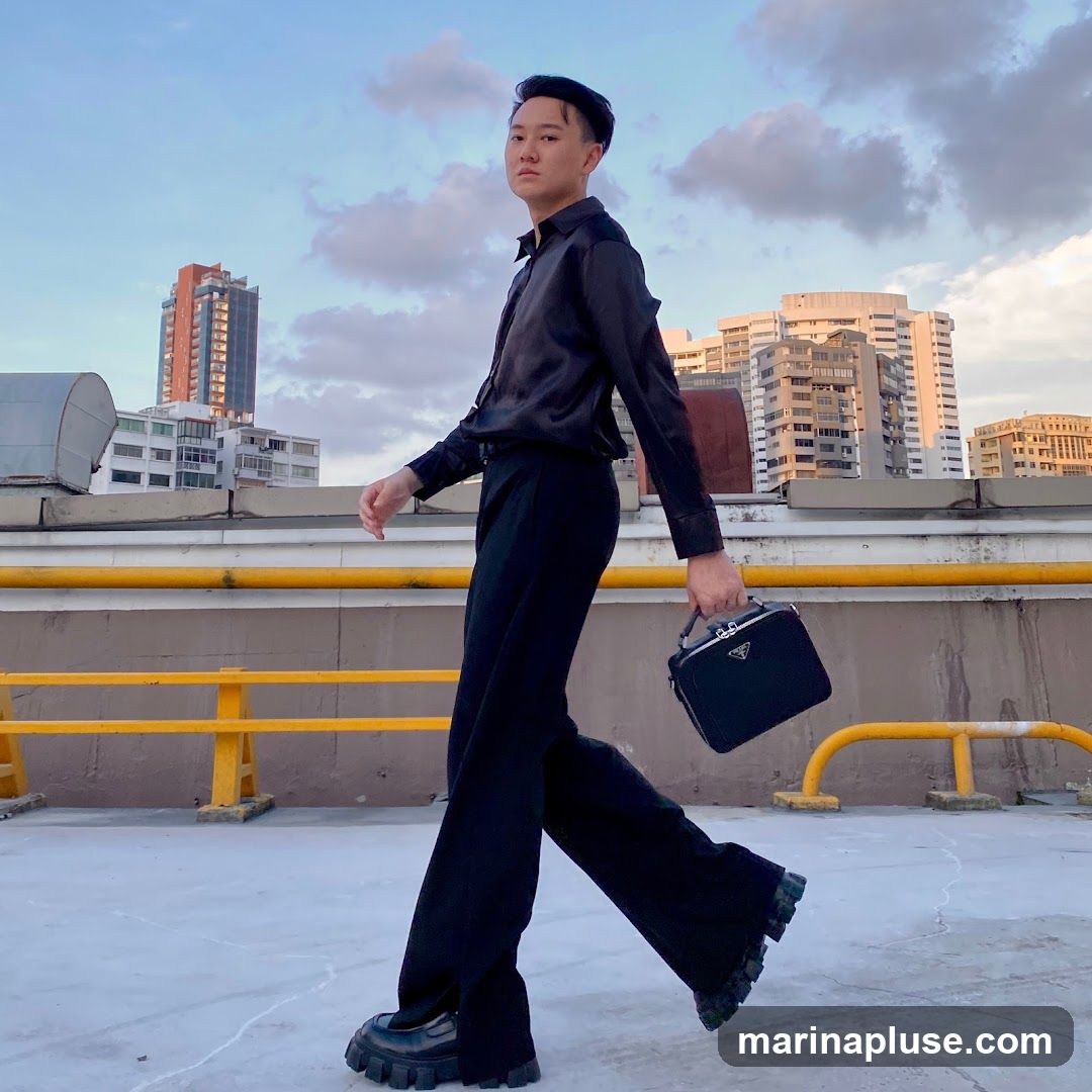 A person dressed in a black satin shirt, black wide-leg pants, and chunky black shoes walks across a rooftop. They are carrying a black handbag with a silver Prada logo. In the background, several city buildings are visible against a cloudy sky.