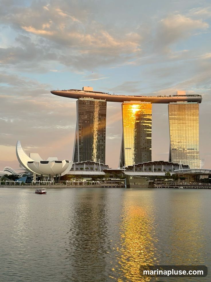 The iconic Marina Bay Sands hotel in Singapore is illuminated by the golden light of the setting sun, with its reflection shimmering on the calm waters of the bay. A small boat navigates the water in the foreground, and the distinctive lotus-shaped ArtScience Museum is visible to the left. The sky is filled with soft, pastel-colored clouds.