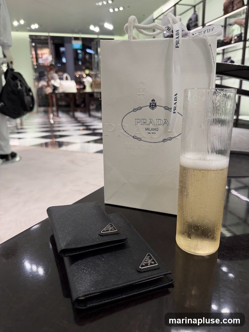 A white Prada shopping bag with embossed logo and ribbon sits on a table next to two black Prada wallets and a glass of champagne. The background shows a blurred interior of a luxury store with checkered flooring and displays of merchandise.