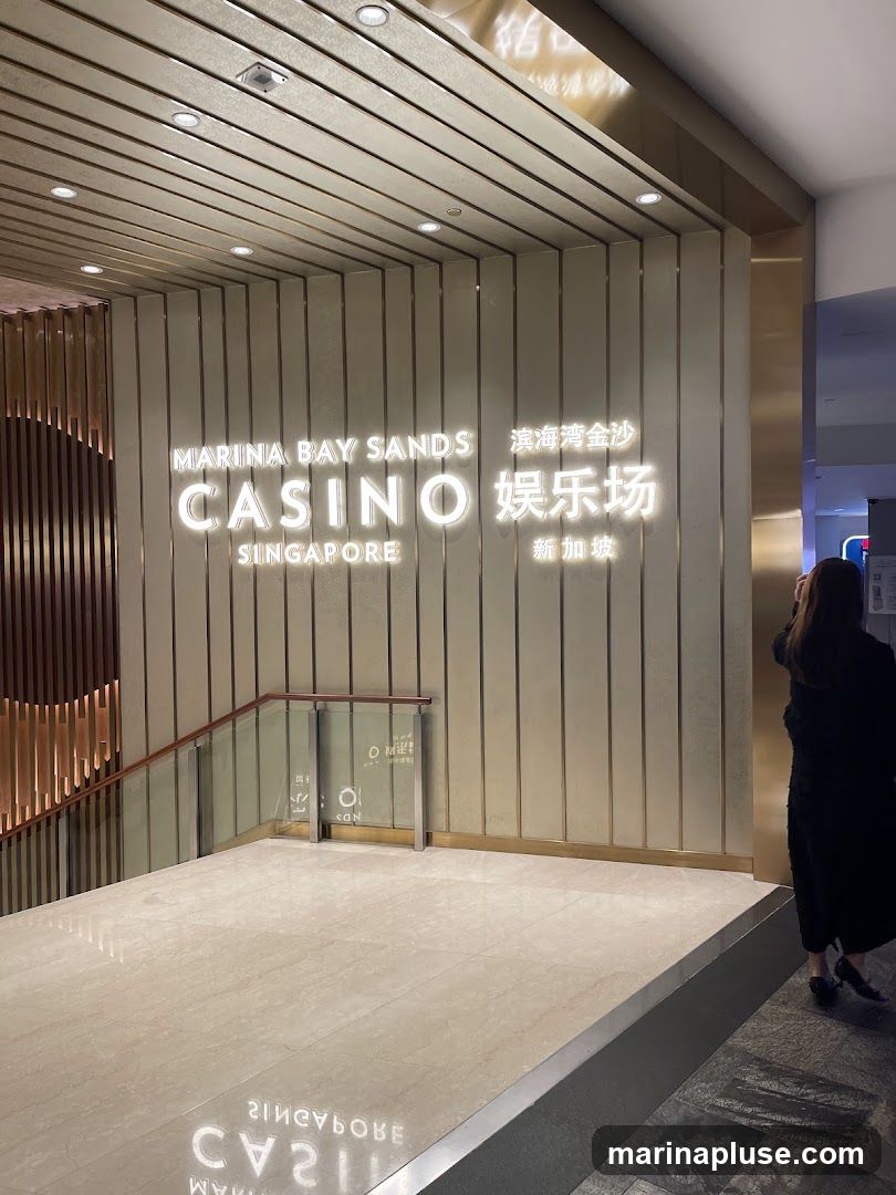 A person walks past a large illuminated sign that reads "MARINA BAY SANDS CASINO SINGAPORE" in English and Chinese. The sign is mounted on a wall with vertical golden panels. A staircase with glass railings is visible to the left, and the floor is made of polished marble, reflecting the signage.