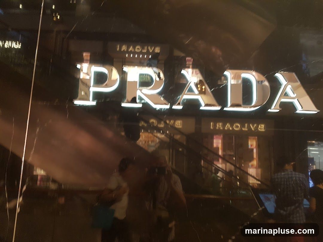 A close-up shot of the Prada logo on a dark, reflective surface. The letters are large, metallic, and illuminated. Below the Prada logo, the word "BULGARI" is visible, also in illuminated letters, but reflected. The storefront appears to be in a shopping mall or high-end retail area, with escalators and other shops visible in the background reflections. Several people are seen in the reflections, some appearing to be shoppers.