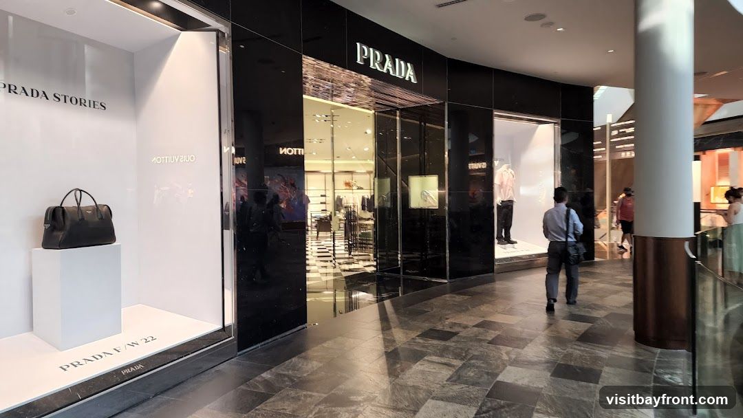 The exterior of a Prada store with large display windows. One window shows a black handbag on a white pedestal with the text "PRADA STORIES" and "PRADA F/W 22" visible. The main entrance features the "PRADA" logo above a glass facade. Inside, clothing and accessories are displayed. People are walking by in the mall corridor.