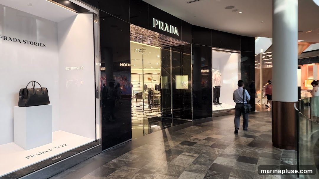 The exterior of a Prada store with large display windows. One window shows a black handbag on a white pedestal with the text "PRADA STORIES" and "PRADA F/W 22" visible. The main entrance features the "PRADA" logo above a glass facade. Inside, clothing and accessories are displayed. People are walking by in the mall corridor.