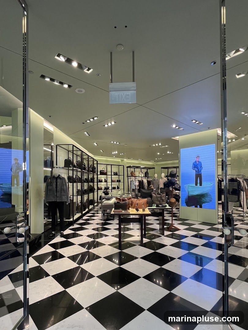 A view from the entrance of a high-end retail store, showcasing a black and white checkered marble floor. The store features displays of handbags, clothing on racks, and mannequins dressed in stylish attire. A large digital screen displays an artistic image of a person in a boat. An illuminated 'EXIT' sign hangs from the ceiling.