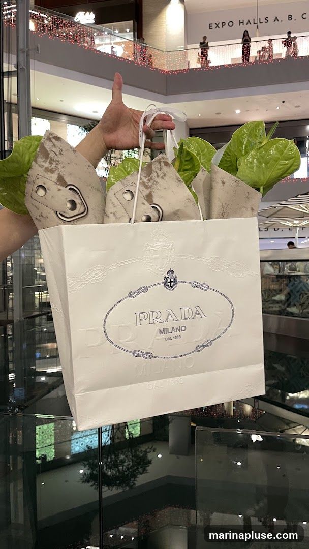 A hand holds a white Prada shopping bag with the brand's logo embossed on it. The bag contains several items wrapped in patterned paper and green leaves. The background appears to be a shopping mall with escalators and decorative lights.