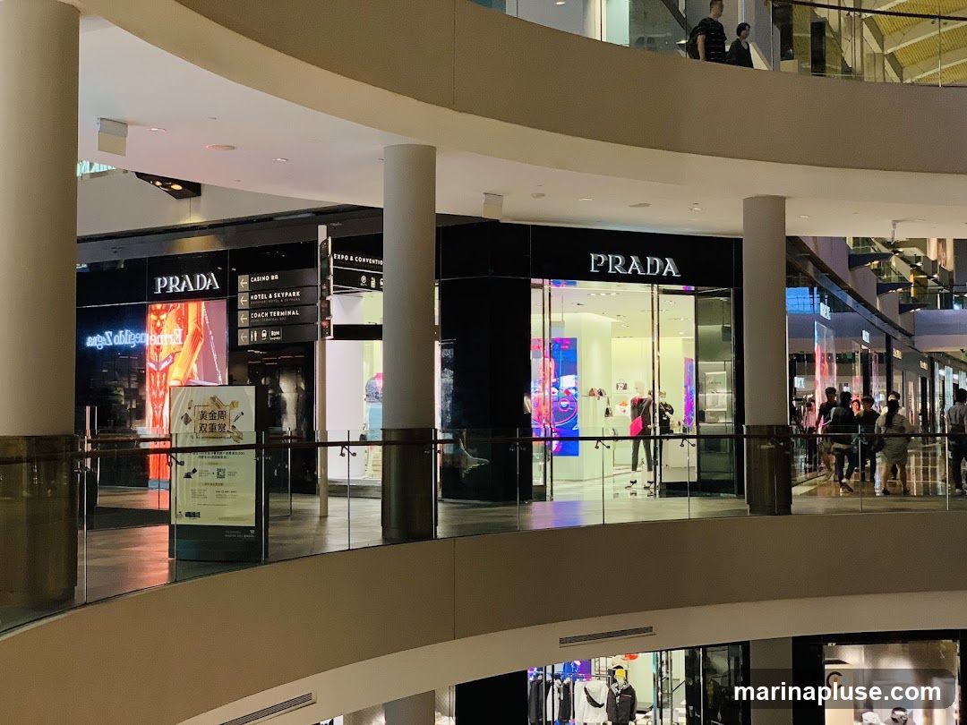 The image shows the exterior of a Prada store within a shopping mall. The store has a large glass facade with the Prada logo prominently displayed above the entrance. Inside, mannequins are visible displaying merchandise. To the left of the Prada store, another storefront with a similar black facade and illuminated displays is visible, also featuring the Prada logo. Signage for