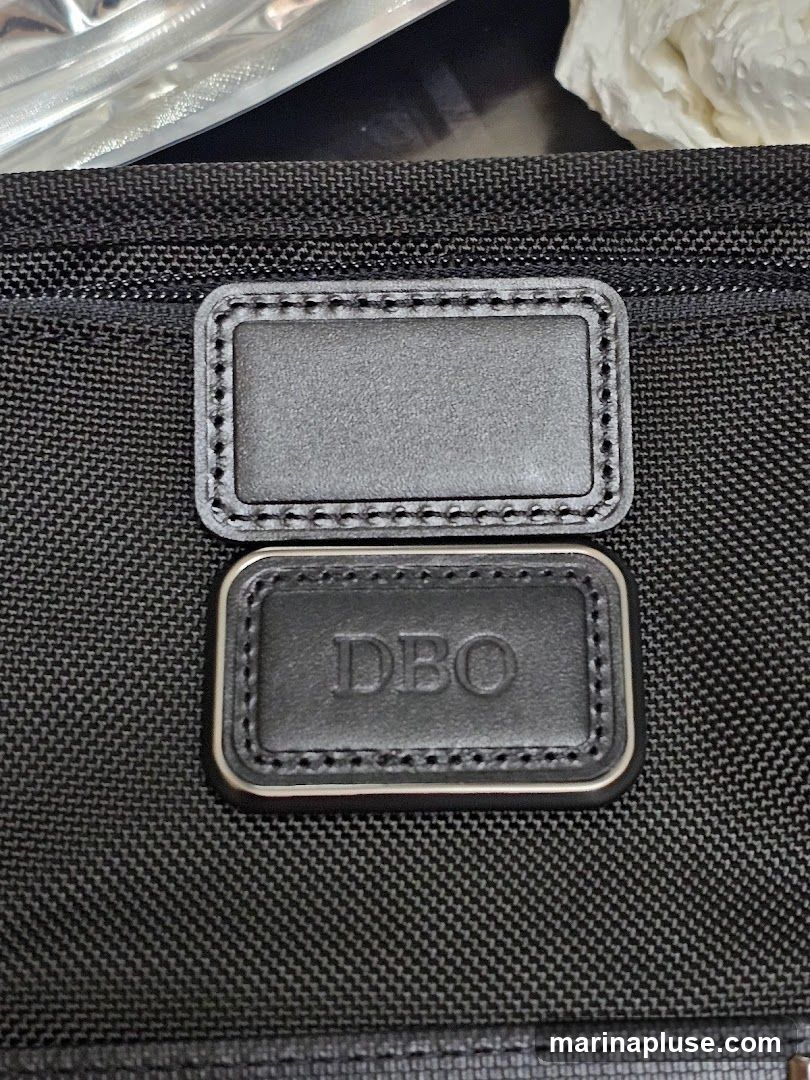 A close-up shot shows two rectangular leather patches on a black textured fabric. The top patch is blank, while the bottom patch has the embossed letters "DBO" in the center. The patches are stitched with a contrasting thread and have a metallic trim around the bottom one.
