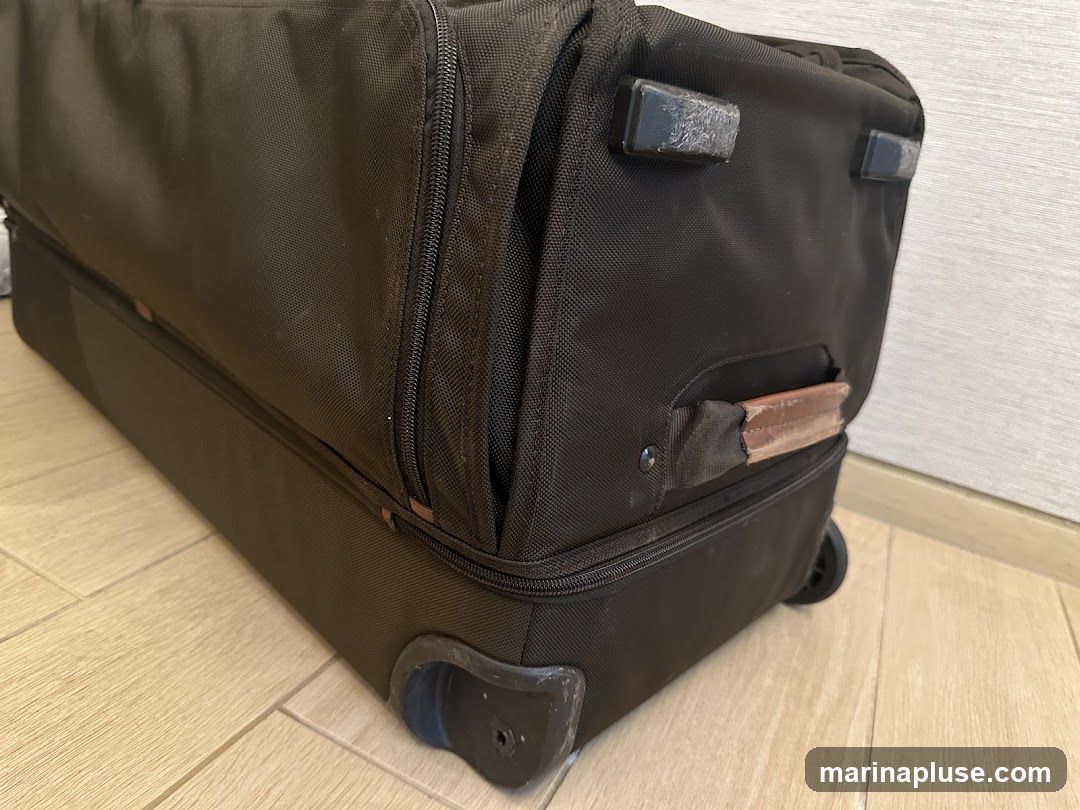 A close-up, low-angle shot of a black rolling suitcase. The suitcase is made of fabric and has zippers and a leather handle. It is resting on a light-colored wooden floor. The suitcase appears to be well-used, with some scuff marks visible on the wheels and corners.