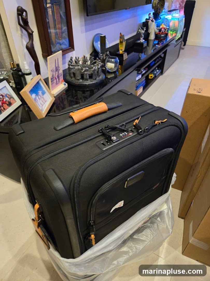 A black suitcase with orange accents is placed in a clear plastic bag. The suitcase has a combination lock and a retractable handle. In the background, a black entertainment center is visible, adorned with various decorative items including framed photos, sculptures, and aquariums.