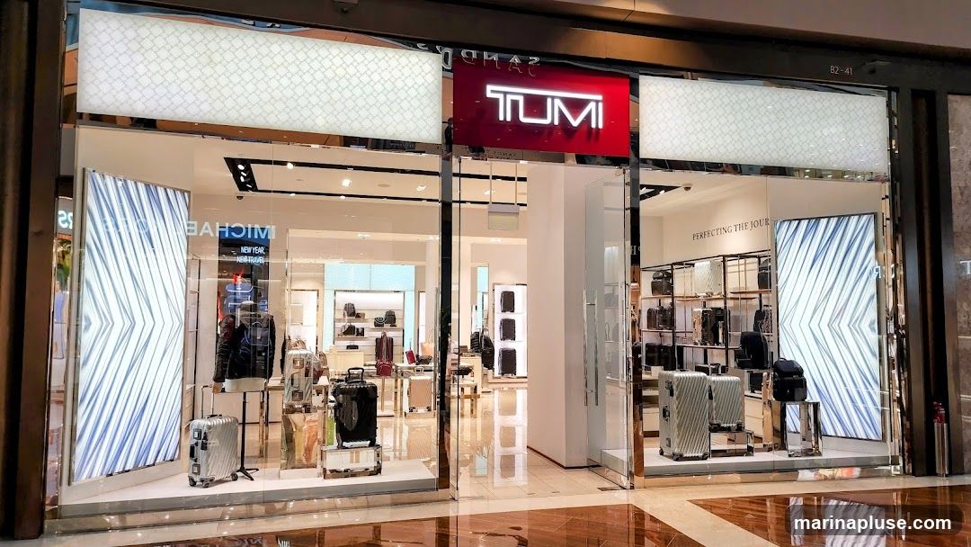 The exterior of a Tumi store, featuring a prominent red sign with the Tumi logo. The storefront displays various luggage and bags, with illuminated panels on either side of the entrance.