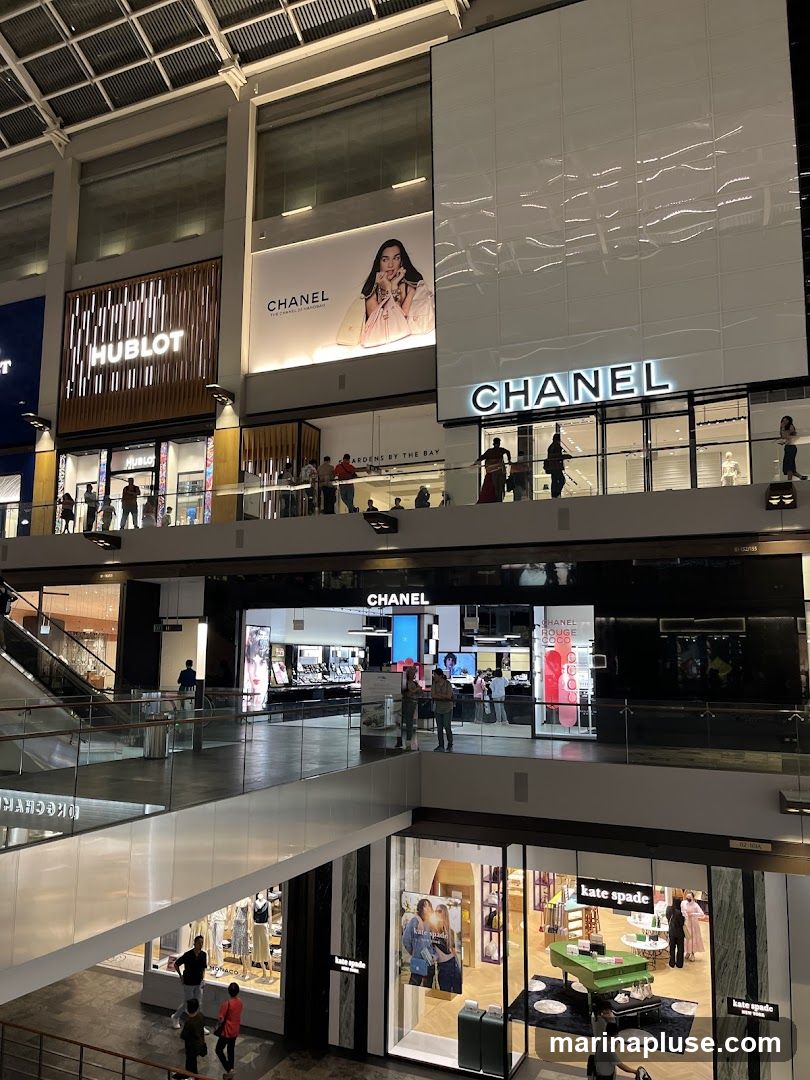 An interior view of a multi-level shopping mall featuring high-end brands. On the upper levels, prominent signage for HUBLOT and CHANEL is visible, along with large advertisements. Further down, another CHANEL store is seen, and a KATE SPADE store with a distinctive green piano display. People are seen browsing and walking through the mall.