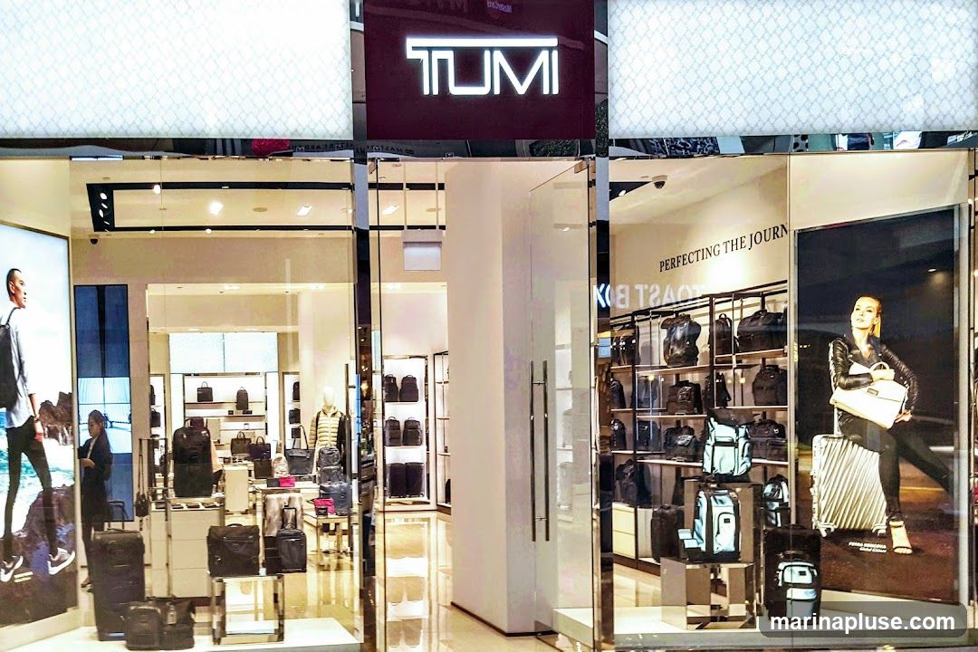 The exterior of a Tumi store, featuring a prominent illuminated Tumi logo above the entrance. The storefront displays various bags and luggage on shelves and stands, with large advertisements showcasing models with Tumi products. The interior is visible through the glass facade, revealing a well-lit retail space with more merchandise.