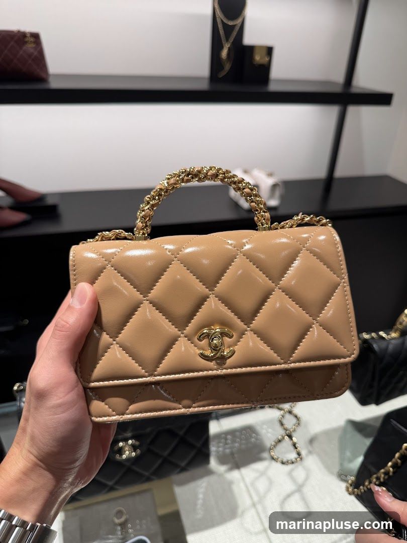 A close-up shot of a beige Chanel handbag with a quilted pattern and a gold chain handle, held by a person's hand. In the background, other Chanel bags and display shelves with jewelry are visible.