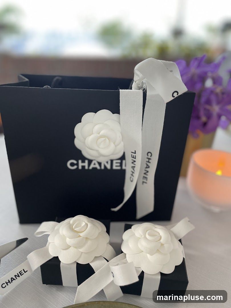 A black Chanel shopping bag with a white camellia flower and ribbon is displayed on a white tablecloth. Two small black boxes, also adorned with white camellia flowers and ribbons, are in front of the bag. A lit candle and purple flowers are visible in the blurred background.