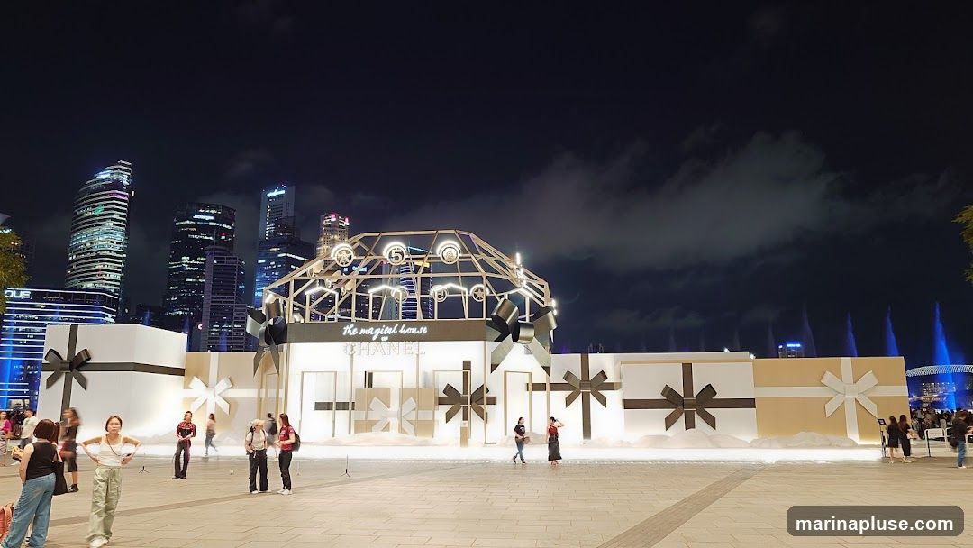 An outdoor nighttime scene featuring a large-scale installation designed to look like oversized gift boxes. The installation has a prominent Chanel logo and the text "the magical house". People are seen walking around the plaza in front of the display. In the background, city skyscrapers are illuminated against the night sky, and a water fountain show is visible on the right.