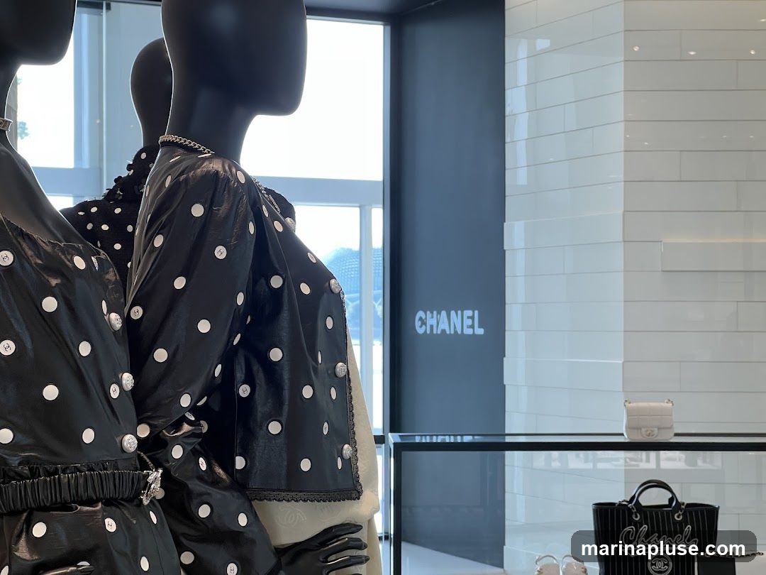 Mannequins dressed in black and white polka dot outfits are displayed in a store. A Chanel logo is visible on the wall. A white handbag and a black tote bag with the Chanel logo are displayed on a shelf.