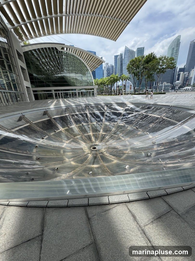 A wide-angle shot captures a modern architectural complex with a large, circular, dry fountain in the foreground. The fountain's metallic surface reflects the sky and surrounding structures. Behind the fountain, a building with a curved glass facade and a distinctive slatted roof is visible. In the background, a dense city skyline with numerous skyscrapers stands under a partly cloudy sky. Palm trees line a walkway leading towards the city. Two people are seen walking in the distance.