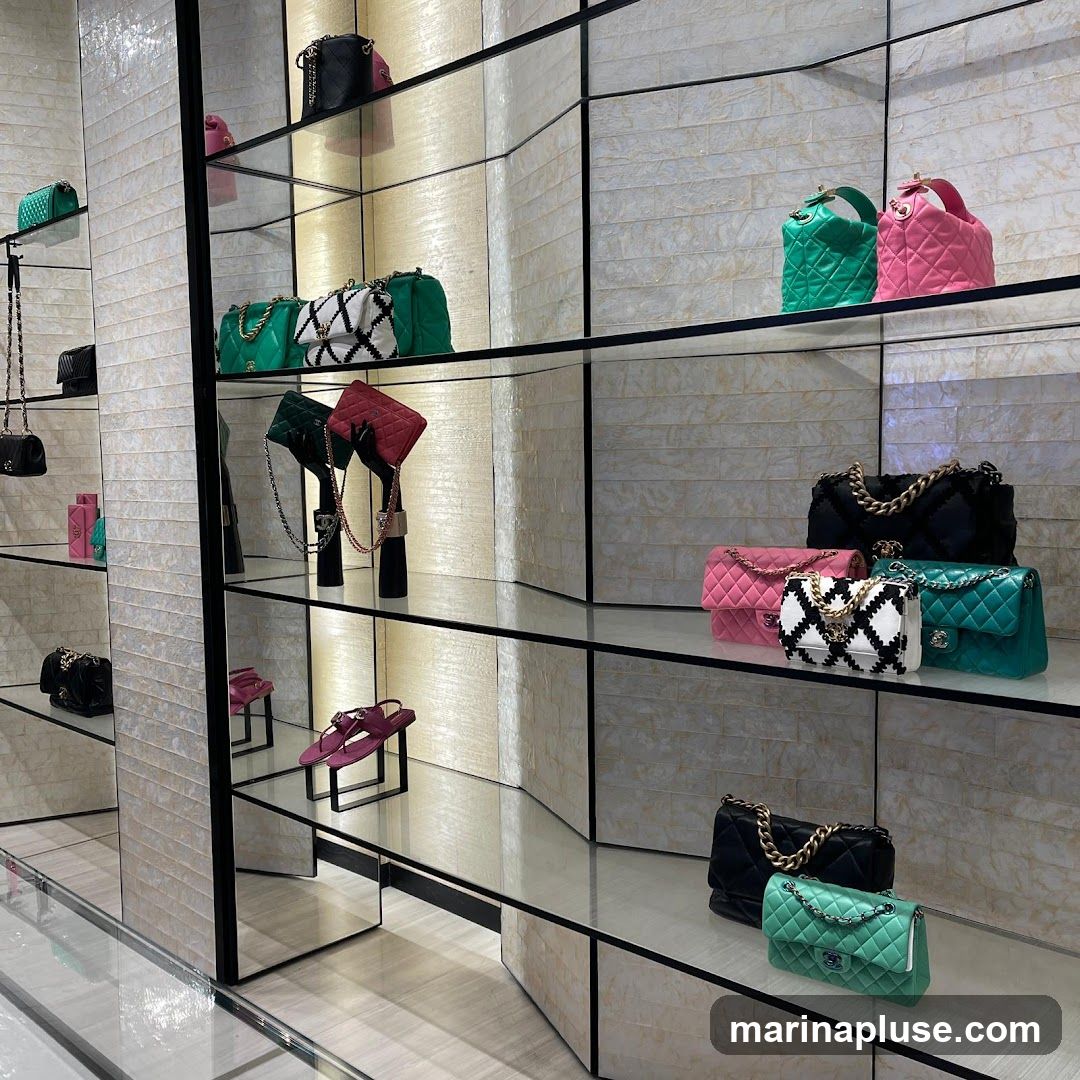 A retail display featuring various handbags and a pair of sandals on glass shelves. The handbags are in different colors including green, pink, black, and white with black patterns. Some are quilted, while others have a diamond-stitched pattern. The sandals are a deep pink color. The shelves are illuminated from behind, creating a soft glow on the textured wall panels.