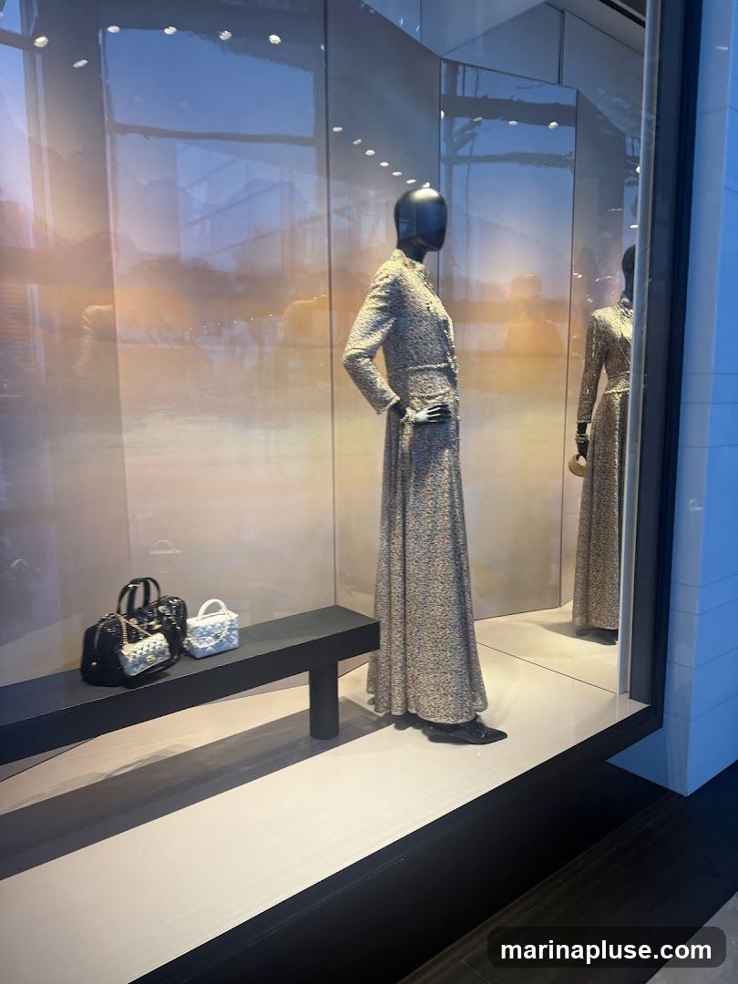 A fashion display window features mannequins dressed in elegant, long floral print dresses. A black bench displays two handbags, one black and one light blue, both with chain straps. The background is reflective, showing a blurred outdoor scene and interior lighting.