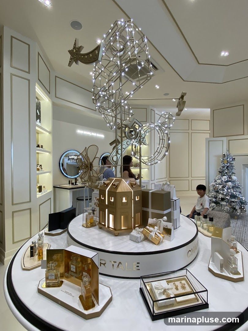 A festive holiday display inside a Chanel store, featuring a large, illuminated sculpture adorned with lights, a miniature golden house, gift-wrapped boxes, and various Chanel perfumes and gift sets. A Christmas tree decorated with white lights is visible in the background.