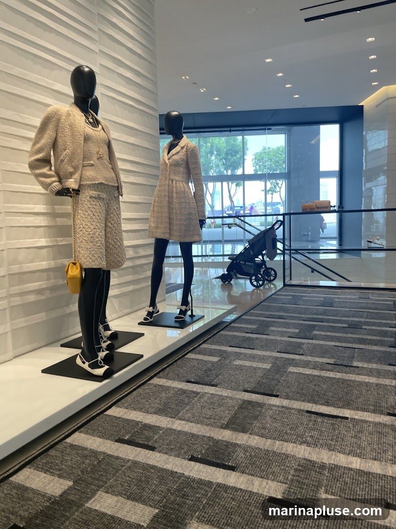 Mannequins dressed in Chanel tweed outfits are displayed in a modern, well-lit store. One mannequin wears a skirt suit with a yellow handbag, while another wears a dress. A baby stroller is visible in the background, along with large windows offering a view of the outdoors. The store features a textured wall and a patterned carpet.
