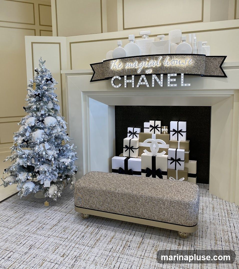 A festive Chanel-themed display features a flocked Christmas tree adorned with black bows and white ornaments. A fireplace mantel is decorated with illuminated Chanel branding and a banner reading "the magical house of CHANEL." Below the mantel, a stack of gift-wrapped boxes in black, white, and gold is arranged, with a large Chanel logo at the center. A textured ottoman sits in front of the fireplace.
