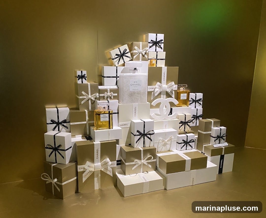 A display of Chanel gift boxes and perfume bottles arranged in a tiered pyramid against a gold background. The boxes are wrapped in white and gold paper with black and white ribbons. Prominently featured are bottles of Chanel No. 5 perfume and a large white Chanel logo.