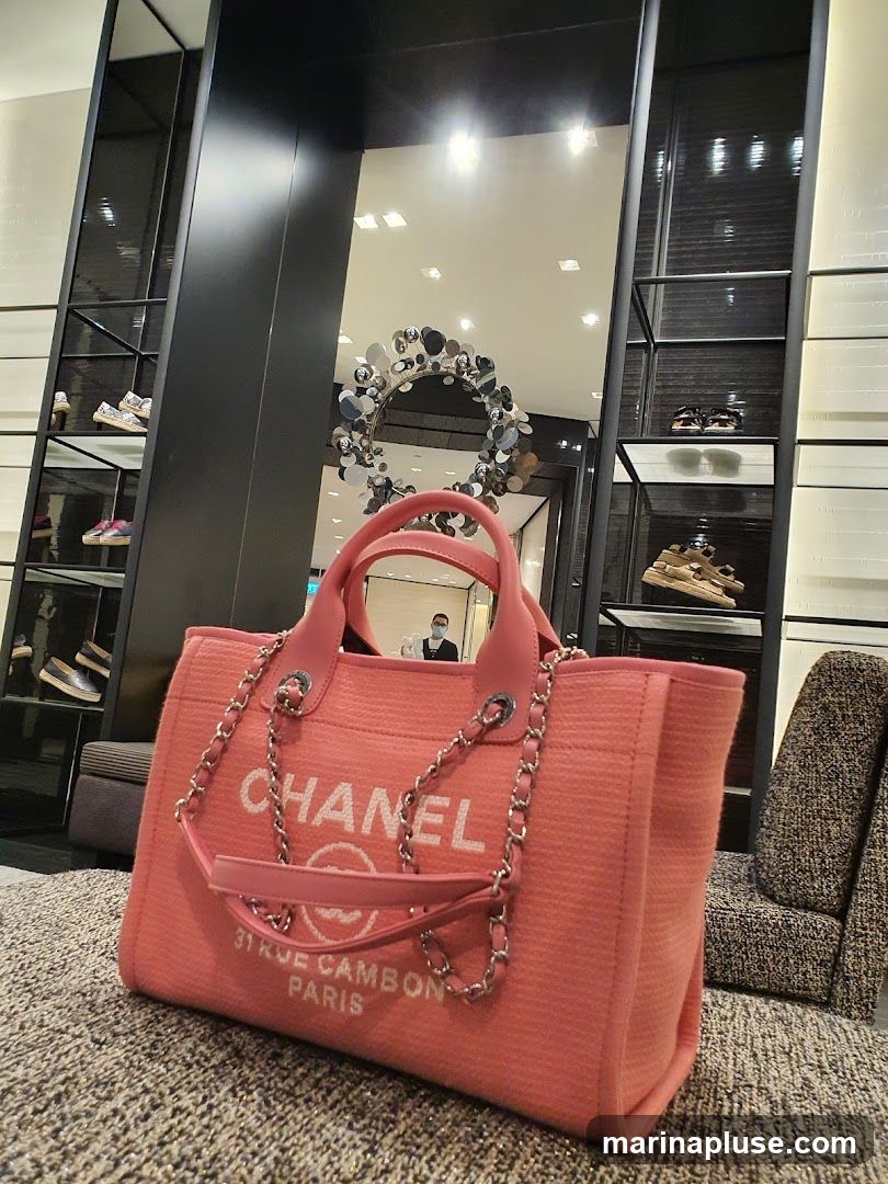 A close-up shot of a pink Chanel tote bag with silver chain straps. The bag features the Chanel logo and "31 RUE CAMBON PARIS" printed in white. In the background, shelves display shoes and accessories, and a circular art piece hangs on the wall. A person wearing a mask is visible in the reflection of a mirror.