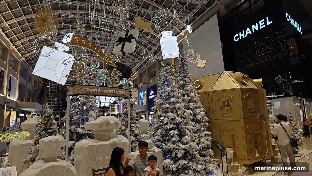 A festive Christmas display featuring large, stylized perfume bottles and gift boxes suspended from the ceiling. Snow-covered Christmas trees and a golden, house-like structure with Chanel branding are prominent. The Chanel store facade with its iconic logo is visible in the background. Families are seen enjoying the holiday decorations.