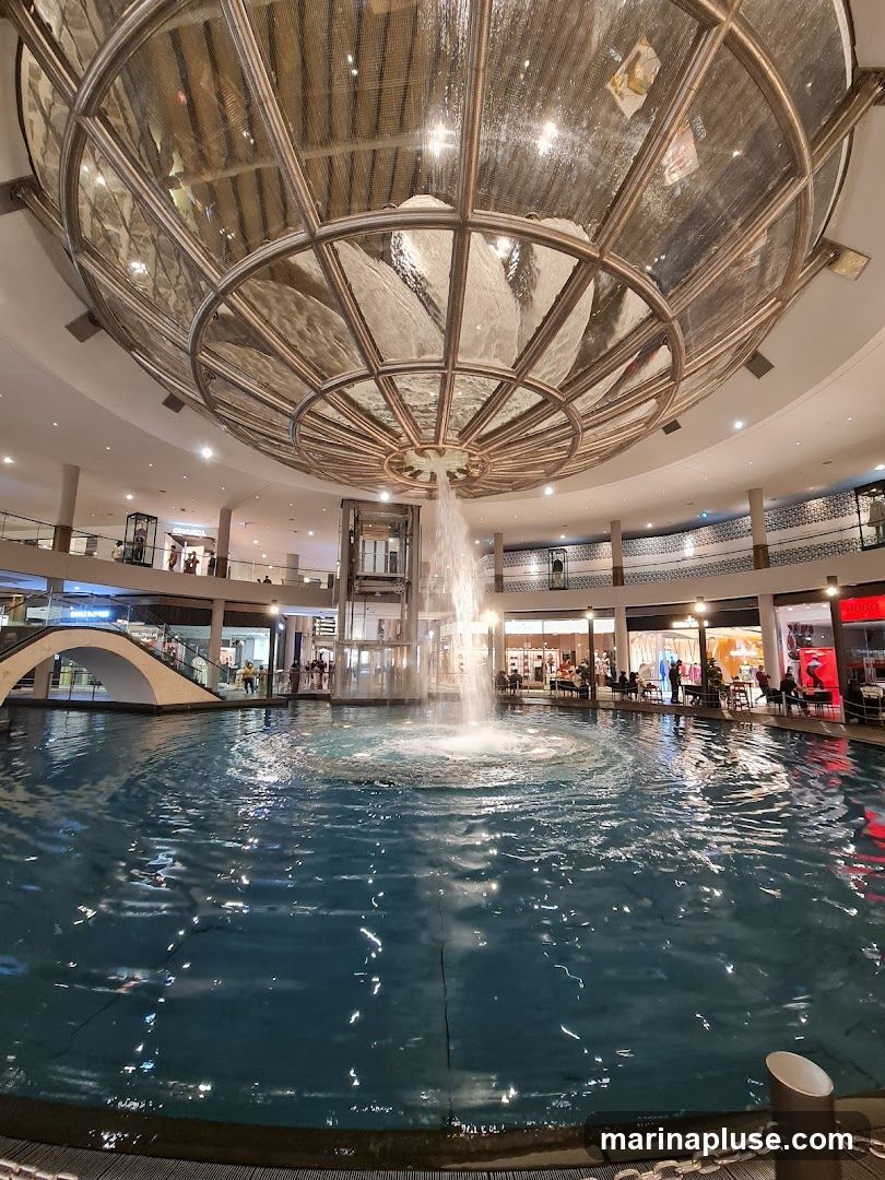 An indoor fountain with water jets is the central feature of a large, multi-level shopping mall. The fountain is surrounded by a pool of water, and shops and escalators are visible on the upper levels. A large, ornate glass dome with metallic accents covers the central area.