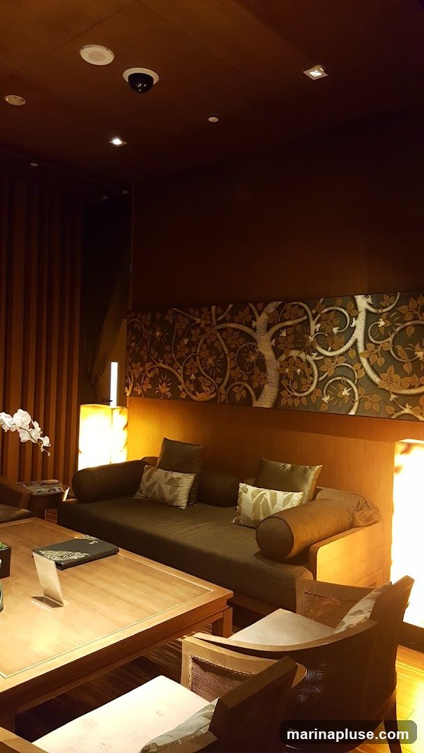 A dimly lit, luxurious seating area with a dark brown sofa adorned with patterned cushions. A wooden coffee table sits in front of the sofa, holding a menu and a small white stand. Behind the sofa, a large decorative panel features an intricate tree motif. To the left, a floor lamp with a warm glow illuminates a white orchid. The room has a sophisticated and relaxing ambiance.