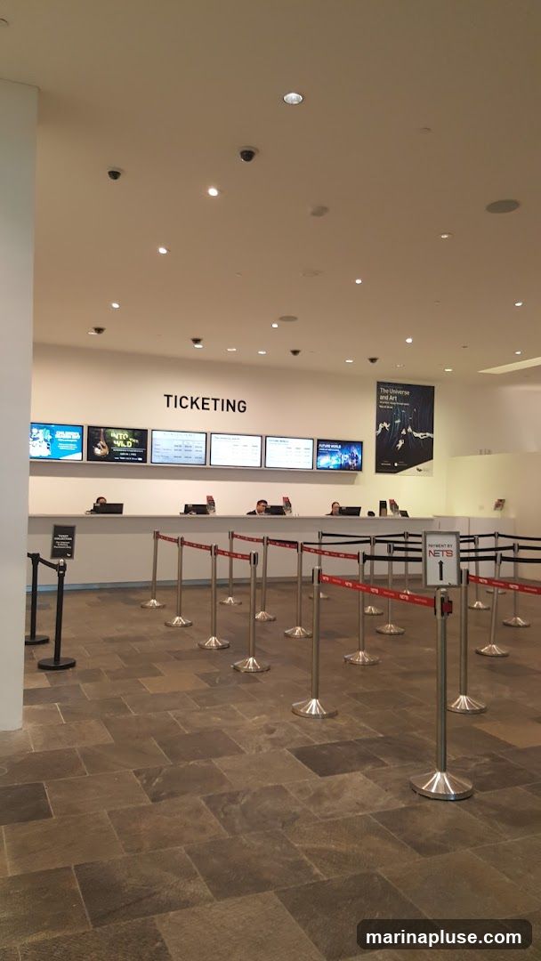 A ticketing counter with multiple screens displaying information and prices. Several employees are visible behind the counter. Stanchions with red ropes guide a queue. A poster titled "The Universe and Art" hangs on the wall.