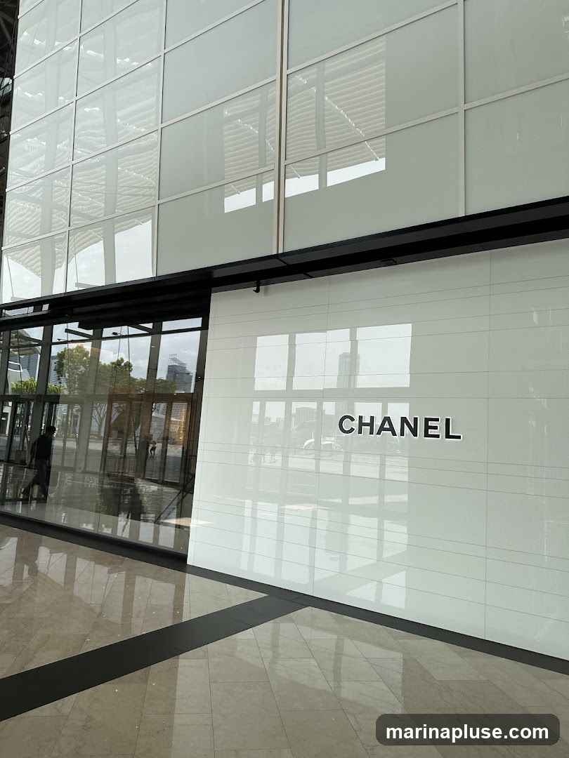 A modern, sleek storefront with the Chanel logo prominently displayed on a white tiled wall. Large glass windows reflect the exterior environment and interior lighting. The floor is a polished marble with dark accent lines.