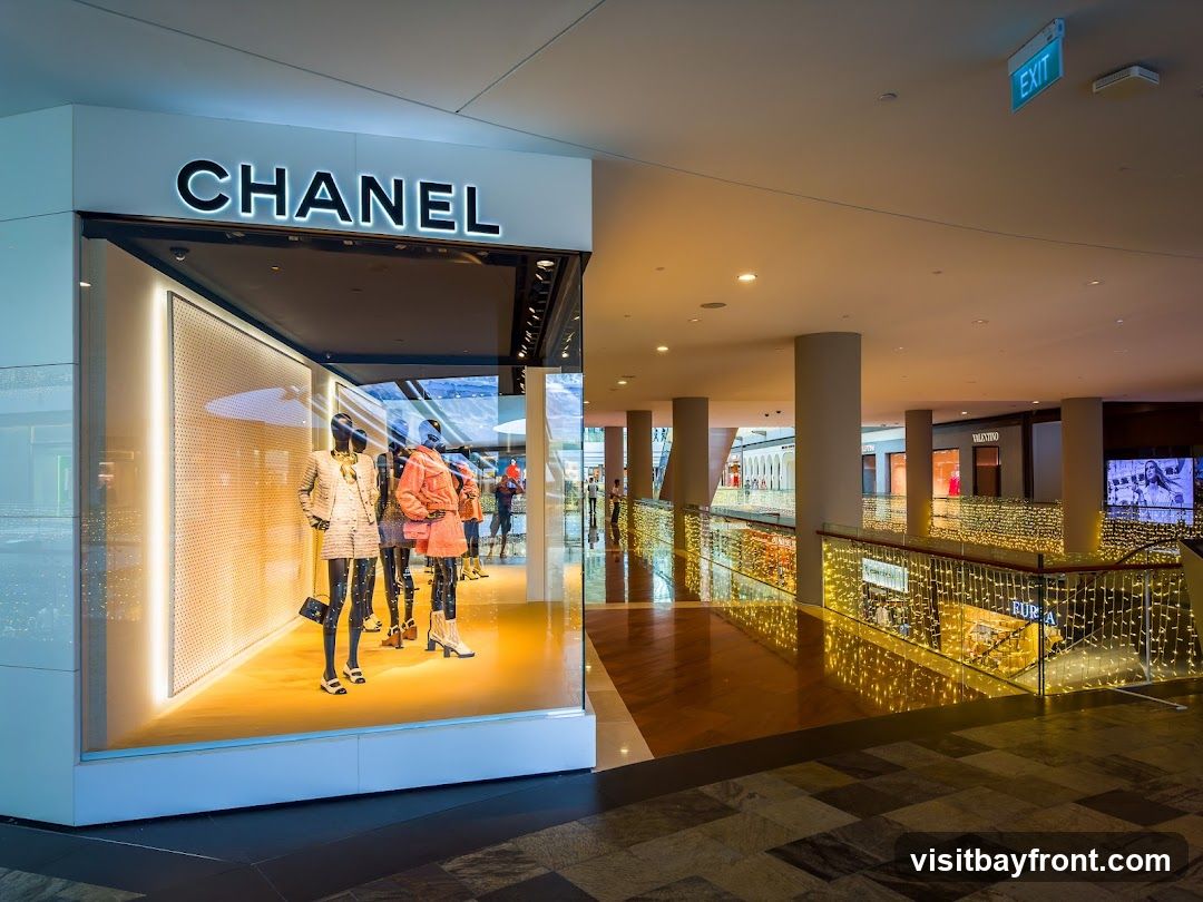 The exterior of a Chanel store with mannequins displaying outfits in the window. The store has a large illuminated sign that reads "CHANEL". The surrounding area appears to be a well-lit shopping mall with decorative lights and other storefronts visible in the distance.