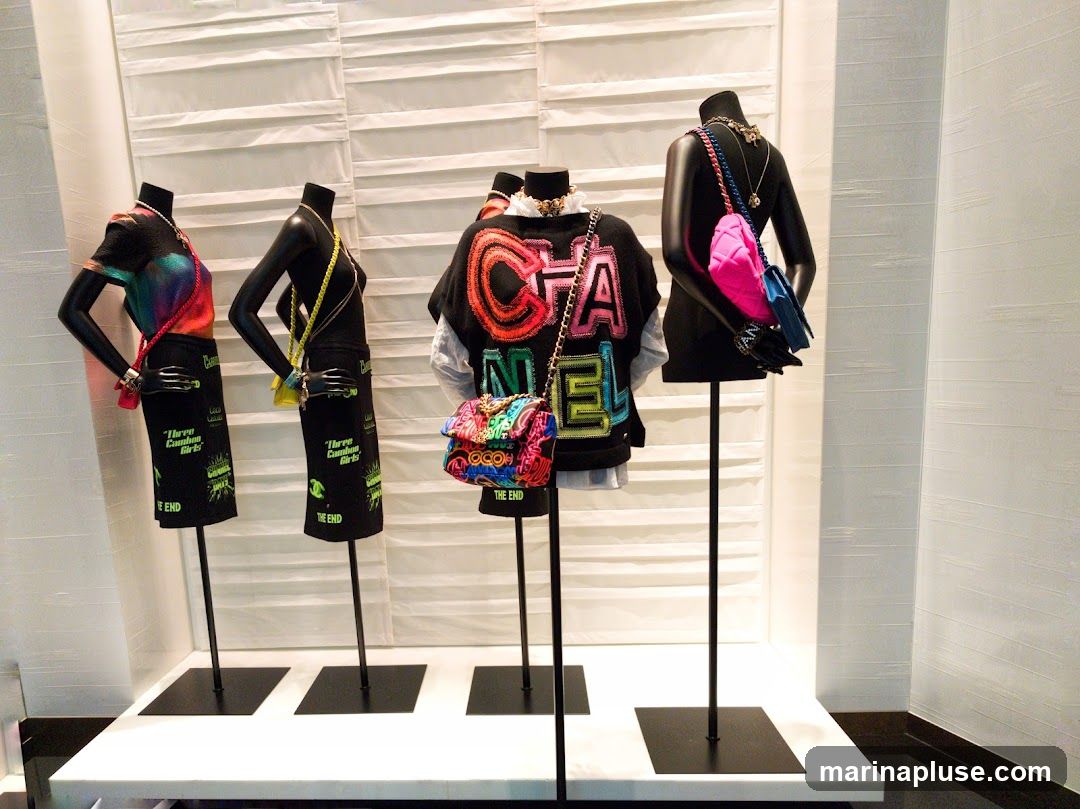 A retail display featuring four mannequins dressed in Chanel clothing and accessories. The mannequins are adorned with colorful handbags, necklaces, and bracelets. The clothing includes skirts with text graphics and a sweatshirt with the Chanel logo in bold, colorful lettering. The background is a textured white wall.