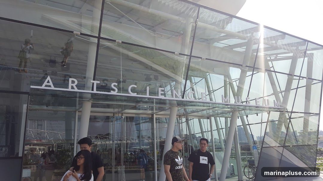 The exterior of the Arts Science Museum with large glass panels and white lettering spelling out "ARTS SCIENCE MUSEUM". People are visible entering and exiting the building.