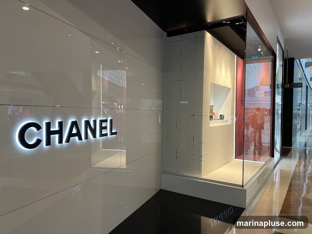 A brightly lit Chanel store facade with the iconic Chanel logo prominently displayed in illuminated black lettering on a white wall. To the right, a display window showcases handbags. The reflection in the glass shows people walking in a mall.