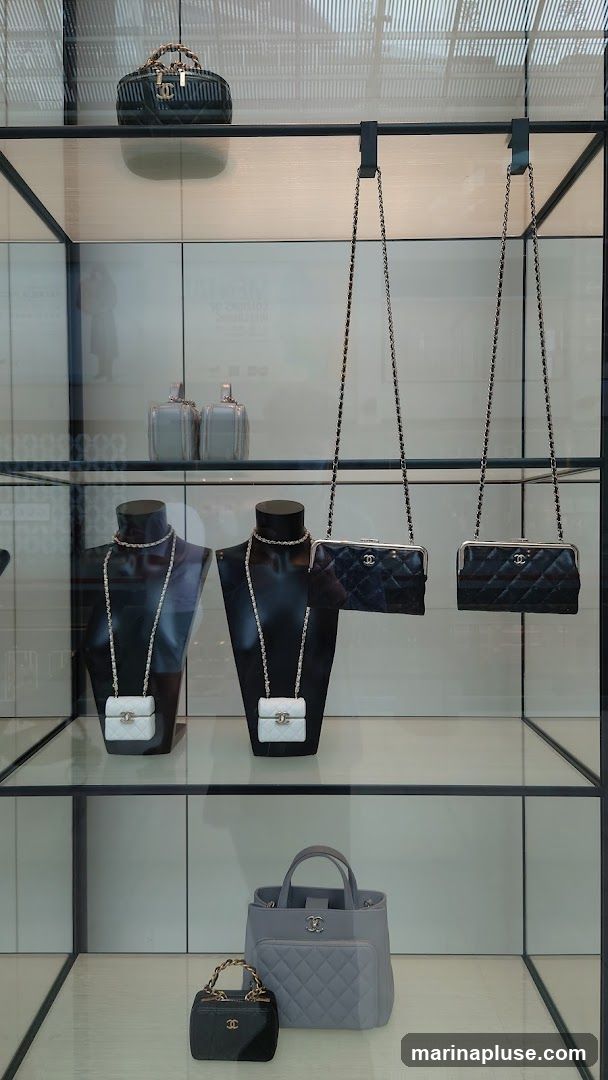 A display window showcases various Chanel handbags. On the top shelf, a dark quilted vanity case with a chain strap and bamboo handle is visible. Below, two black clutches with chain straps hang, and two white mini flap bags are displayed on black busts with chain necklaces. The bottom shelf features a small black vanity case with a chain handle and a larger grey tote bag with a quilted front pocket.