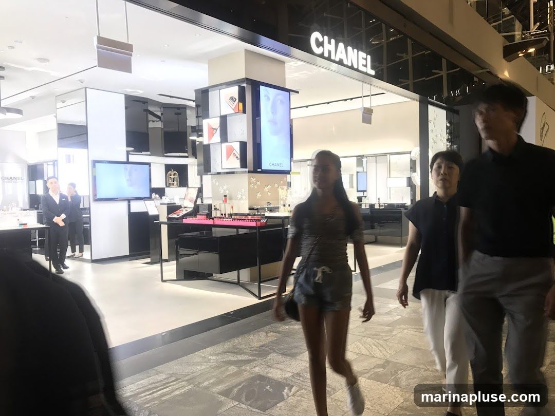 The exterior of a Chanel store with the brand name prominently displayed above the entrance. Inside, displays of makeup and screens are visible. Several people are walking past the storefront.