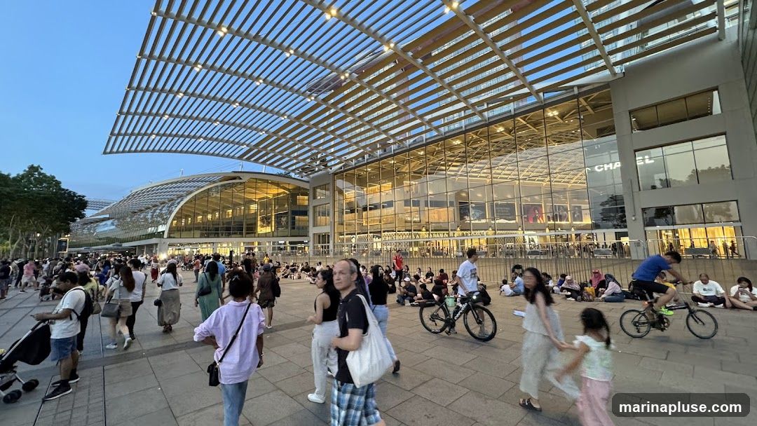 A wide shot of a bustling outdoor plaza in front of a large, modern building with a distinctive curved, slatted roof. Many people are walking, sitting, and cycling in the plaza. The building features extensive glass facades, reflecting the sky and interior lights. Signage is visible on the building, including what appears to be "CHA EL". The overall atmosphere is lively and suggests a popular public space or shopping area.