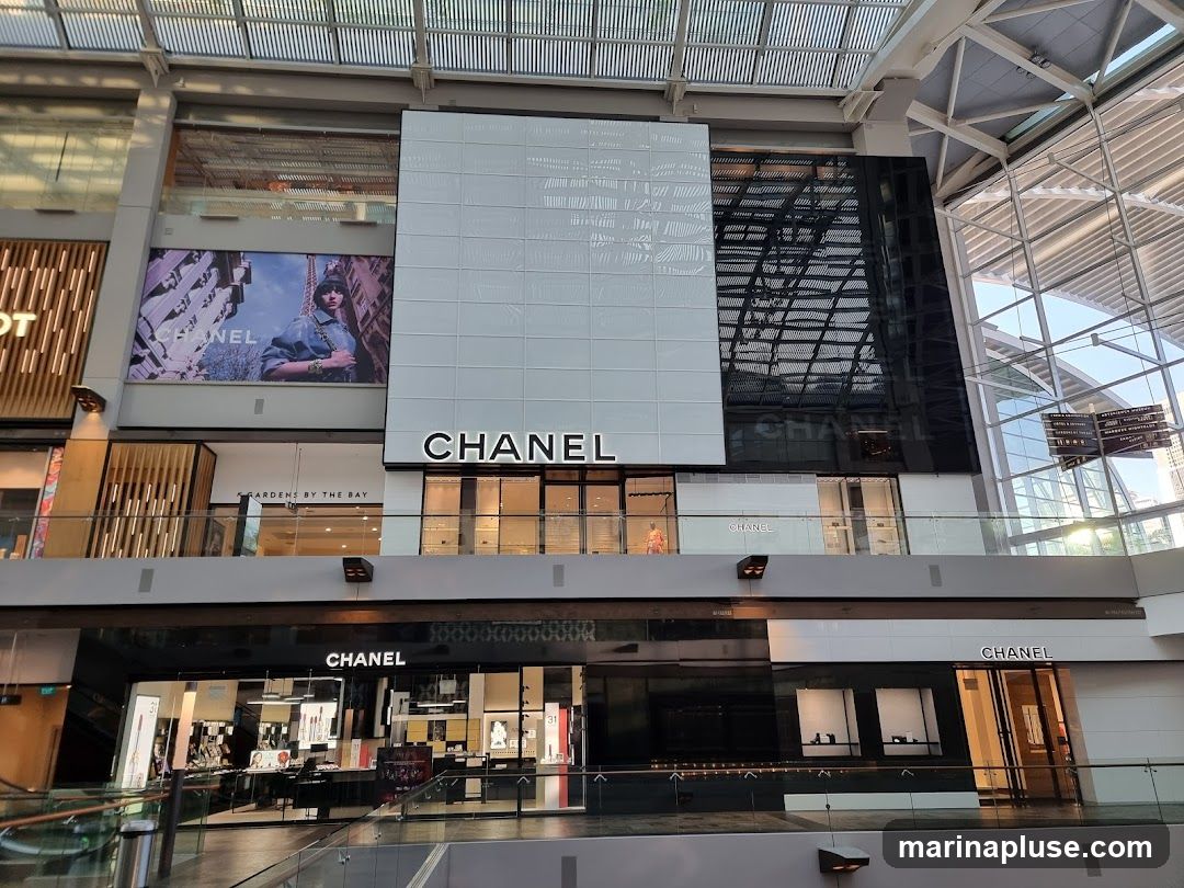 A multi-level Chanel store is visible within a modern shopping mall. The upper level features a large white facade with the Chanel logo, and a large advertisement with a model wearing denim. Below this, a darker facade also displays the Chanel name, with shop windows showcasing merchandise. The lower level also has a Chanel storefront with illuminated displays.