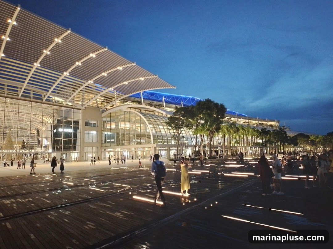 A wide shot of the exterior of Marina Bay Sands, a large complex with a distinctive arched roof structure, illuminated at dusk. People are seen walking on a wooden boardwalk with reflective water features and embedded lights. The building facade features large glass windows and visible signage for 'CHANEL' and 'MARINA BAY SANDS'. Palm trees and other greenery are visible along the building's edge. The sky is a deep blue, indicating twilight.